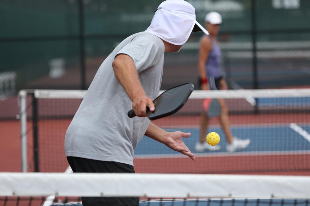 Pickleball-spiller server. I bakgrunnen, på den andre siden av nettet, står motspilleren. Foto.