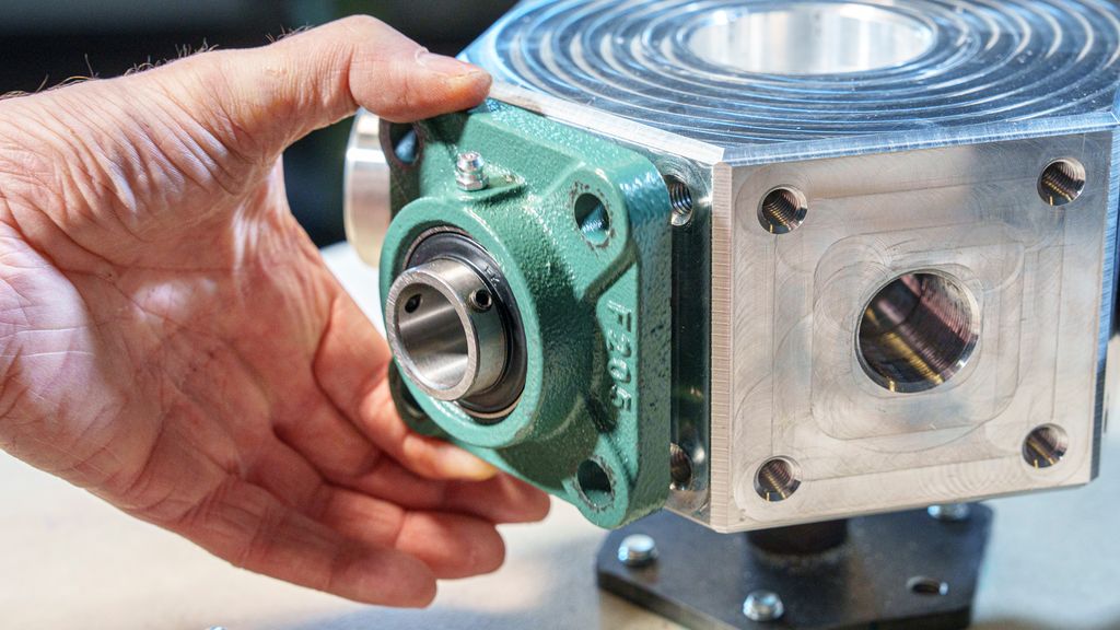 Ei hånd som holder en grønn metallkonstruksjon mot ei sølvfarga festeplate. Foto.