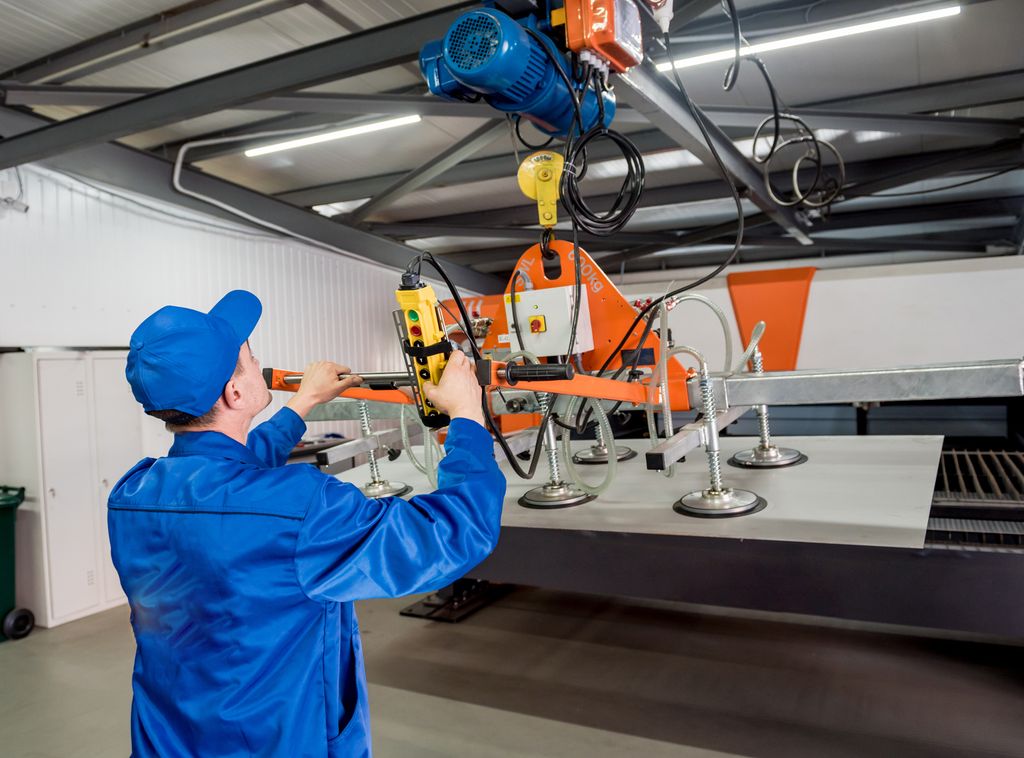 Person i blå arbeidsklær står foran en arbeidsbenk og betjener ei vakuumpumpe. Han holder et betjeningspanel i høyrehanda. Med venstrehanda holder han i et handtak som gjør det mulig å flytte på pumpe og sugekopper. Foto.