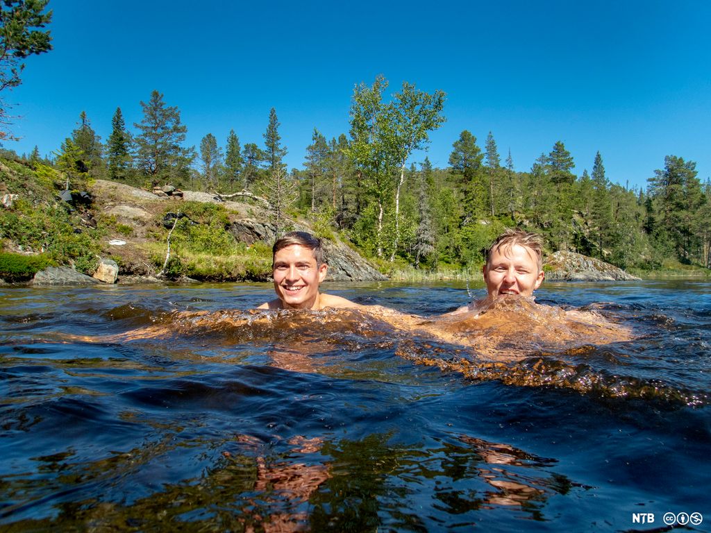 To gutter på rundt 20 år bader i et lite skogsvann. Begge smiler bredt mot kamera. Himmelen er skyfri. Foto.