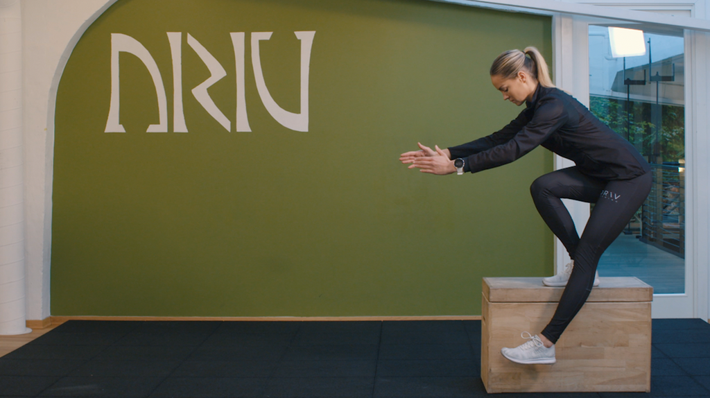 Ei dame står på ett bein på en kasse og senker det andre beinet ned mot gulvet. Foto.