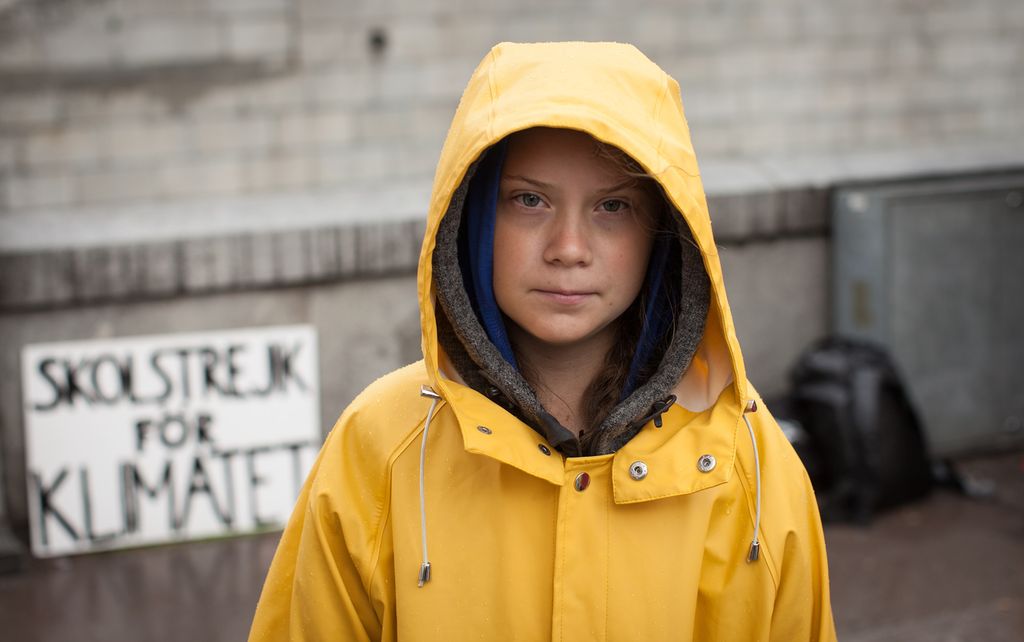 Greta Thunberg, iført ei gul regnjakke. En plakat bak henne har teksten skolestreik for klimaet på svensk. Foto.