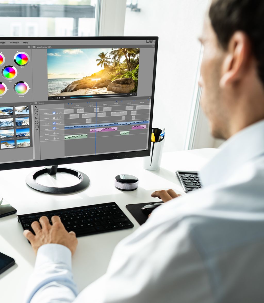 Mann sitter foran en stasjonær datamaskin og redigerer et bilde av en strand. Foto.