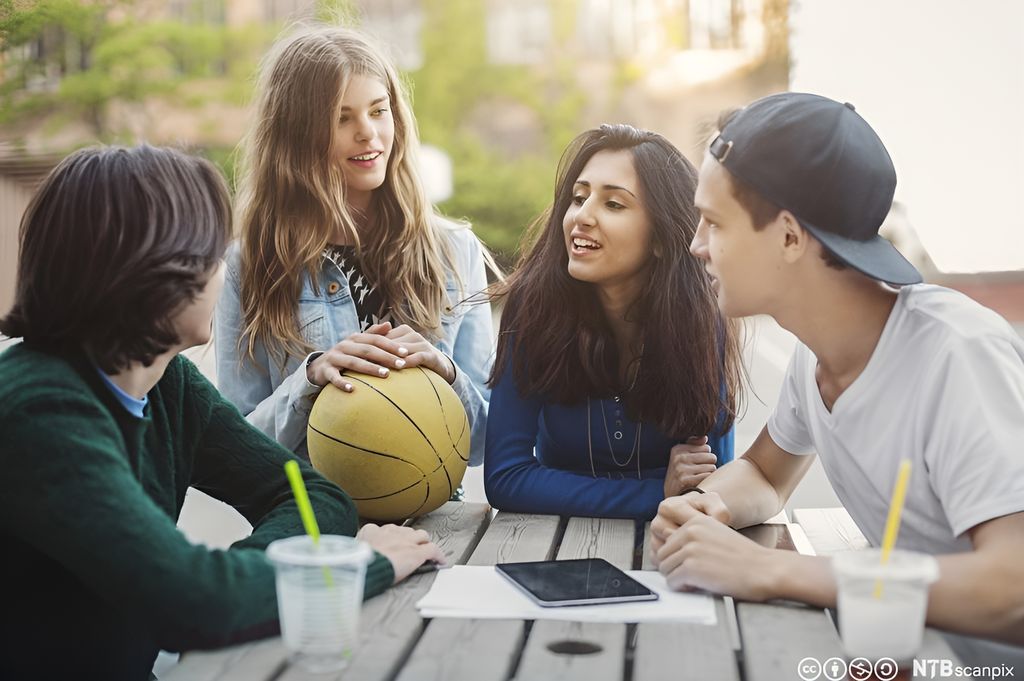 Fire ungdommer sitter rundt et bord i skolegården og prater. Foto.
