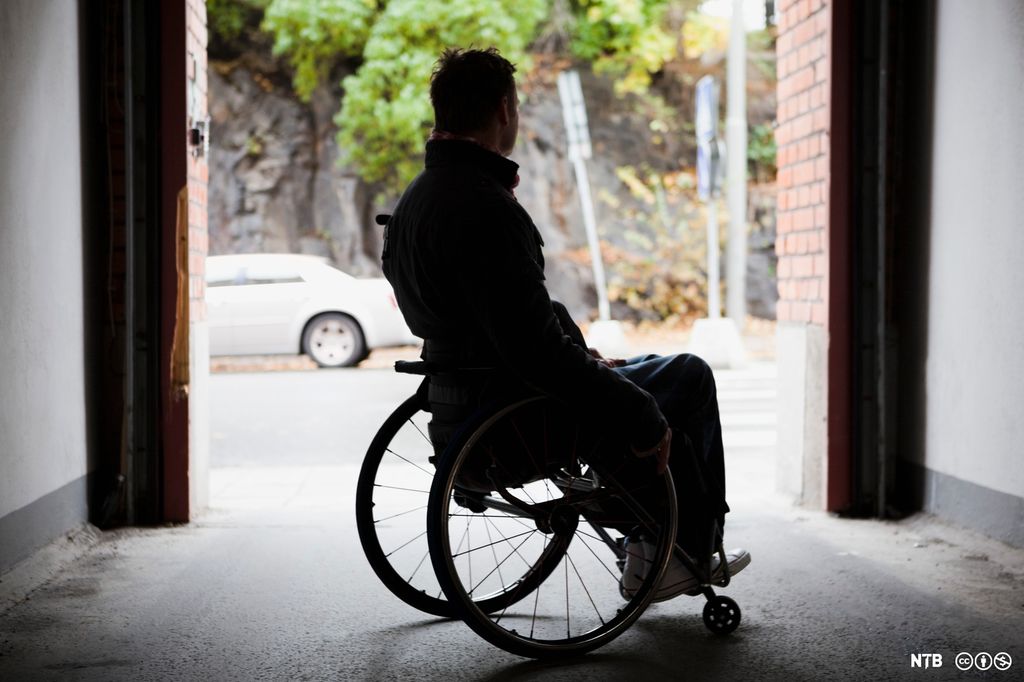 Ein mann sit i rullestol i eit portrom og ser ut på ein veg. Foto.