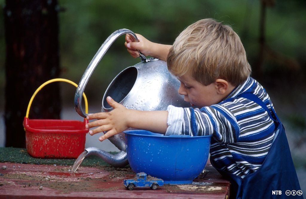 Liten gutt heller ut vann fra en stor kaffekjele på et bord med sand. Foto. 