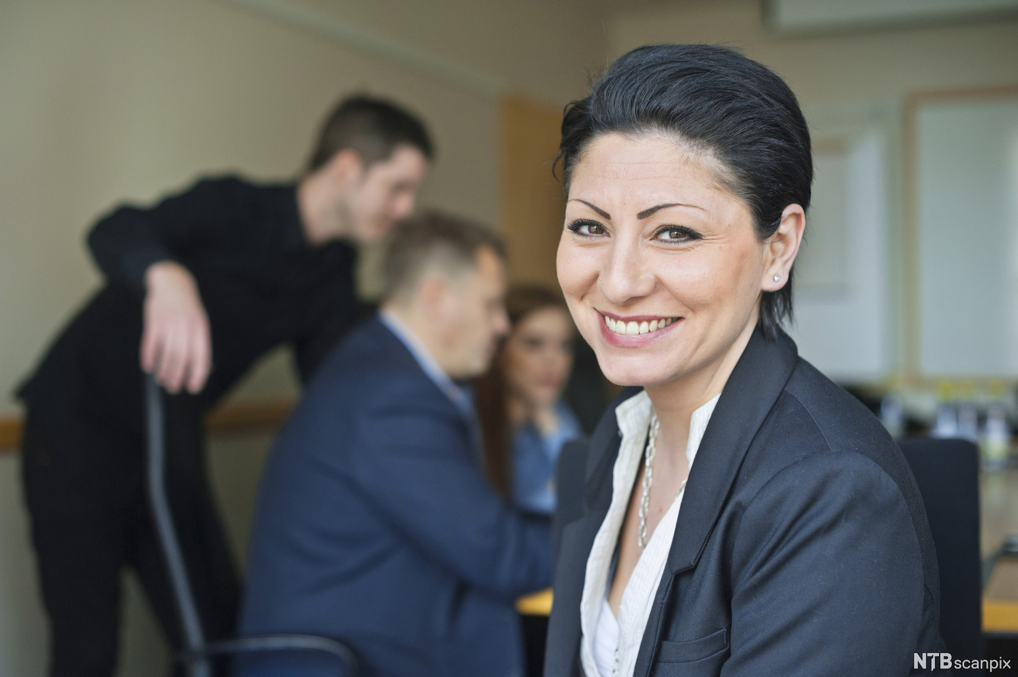 Ei formelt kledd kvinne sitter foran flere medarbeidere i et møte. Hun smiler. Foto.