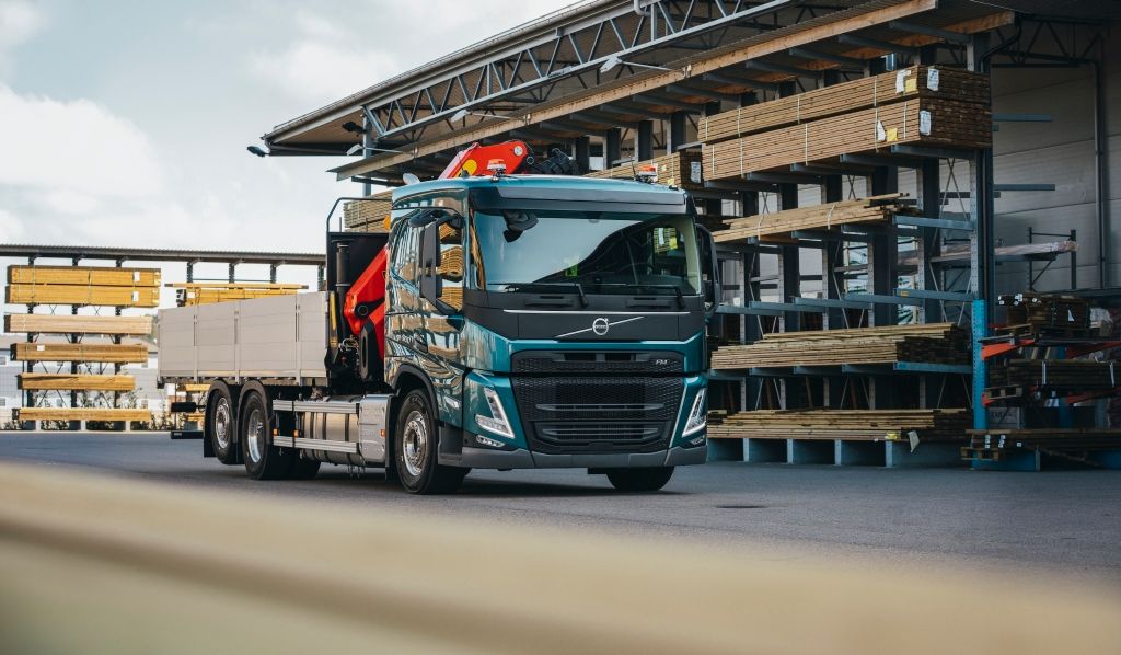 Bildet viser en lastebil med plan og frontmontert kran. Bilen står plassert utafor et lager for byggevarer. Foto.