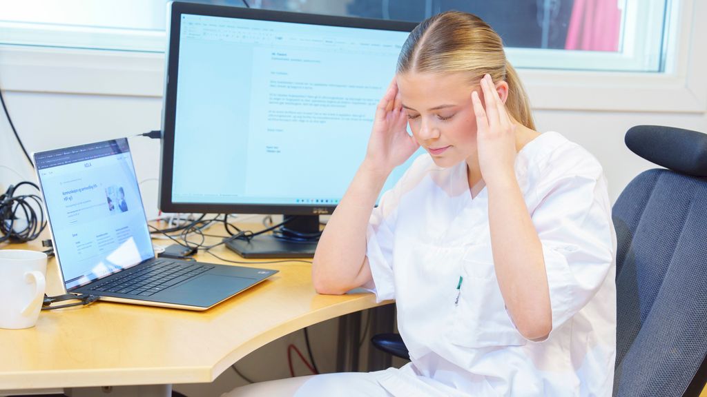 Helseservicearbeider sitter ved kontorpulten sin. Hun lukker øynene og holder hendene mot hodet. Foto.