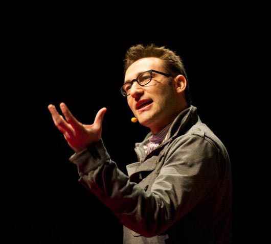 Simon Sinek snakker og gestikulerer. Han har trådløs mikrofon og står i scenebelysning med mørk bakgrunn. Foto.