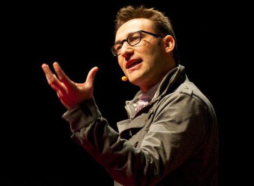 Simon Sinek snakker og gestikulerer. Han har trådløs mikrofon og står i scenebelysning med mørk bakgrunn. Foto.