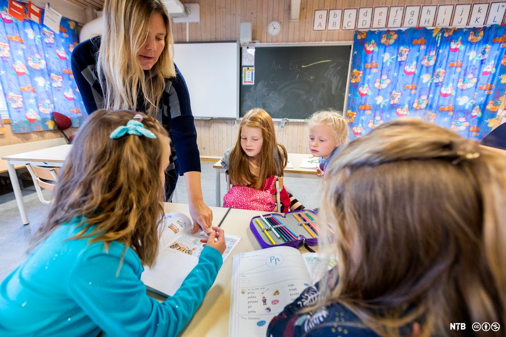 En kvinnelig lærer veileder elever på barneskole. Hun peker i arbeidsboka til den ene eleven, og flere andre elever følger også med. Det er fargerike gardiner i vinduene og bokstavplakater på veggen. Foto.