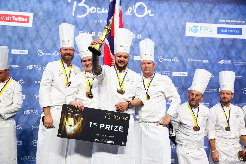 Christian André Pettersen, vinneren av Bocuse d’Or Europe 2020, holder opp pokalen og premiesjekken. I bakgrunnen er det flere andre kokker. Foto.