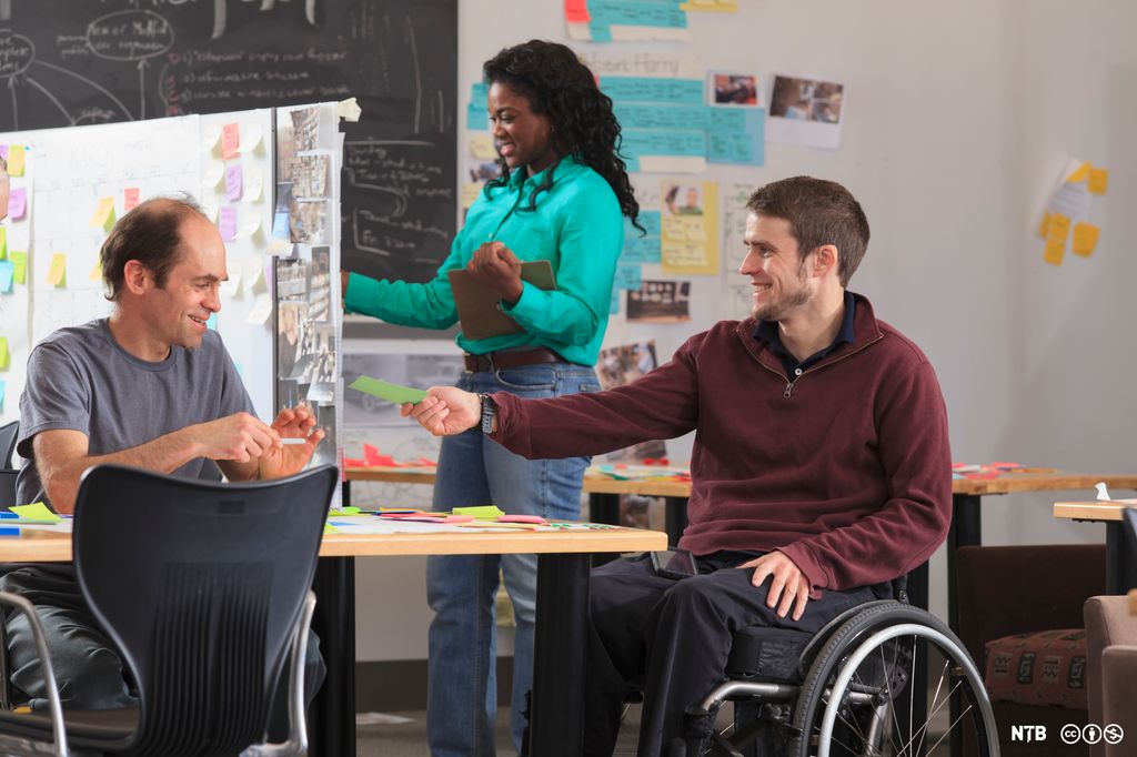 Mann i rullestol gir ein lapp til ein annan mann. I bakgrunnen er ei tavle med mange lappar i ulike fargar. Ei kvinne står og ser på tavla. Foto.