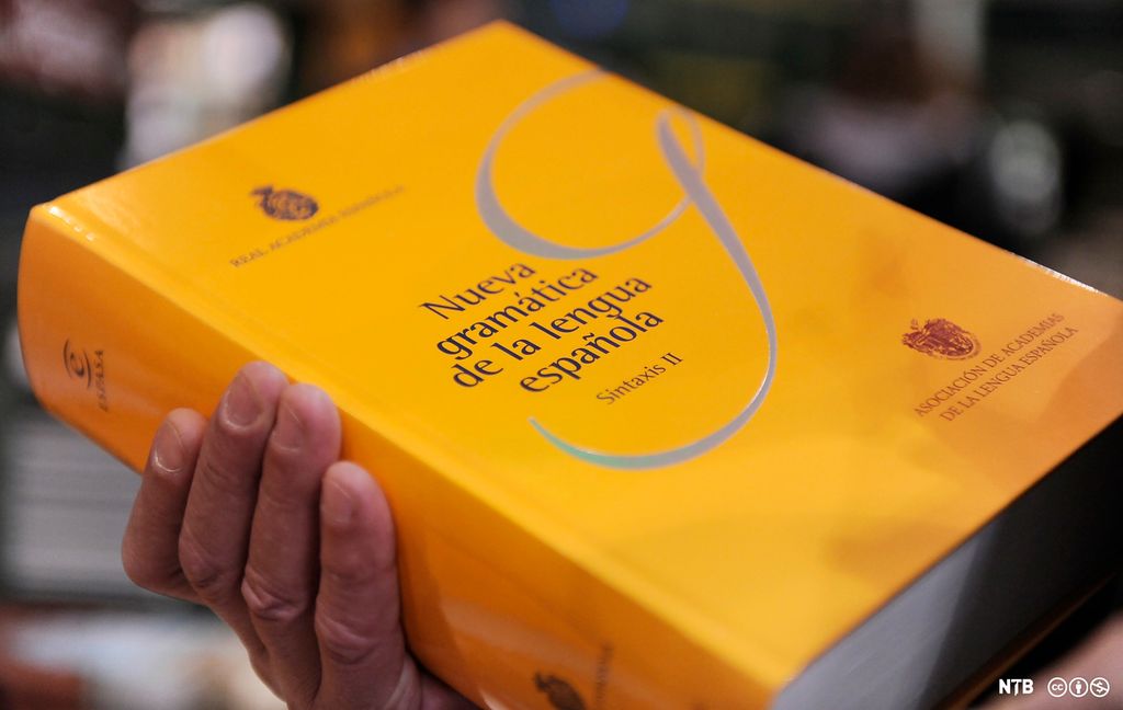 Ei hånd som holder ei gul spansk grammatikkbok. Foto.