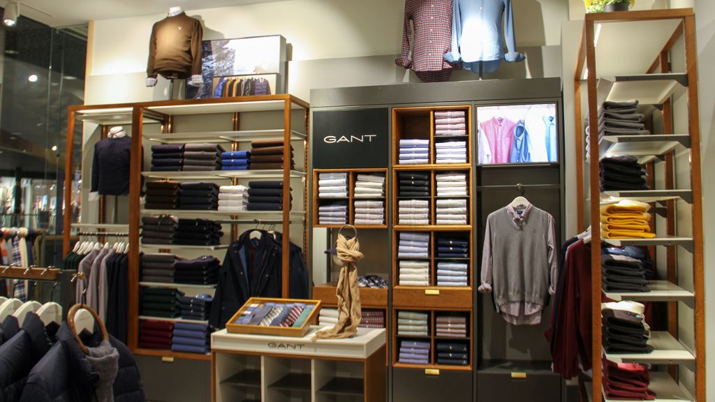 Fra lokalene til en klesbutikk. Utsnittet viser en kombinasjon av hyller med klær, klær på utstillingsbyster, på kleshengere og på utstillingsbord. Logoen til Gant finnes på veggen og på kanten av bordet. Foto.