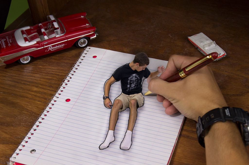 A hand drawing in a notebook, drawing an outline around a boy who sits up on the page of the notebook. There are toy cars on the desk. Photo.