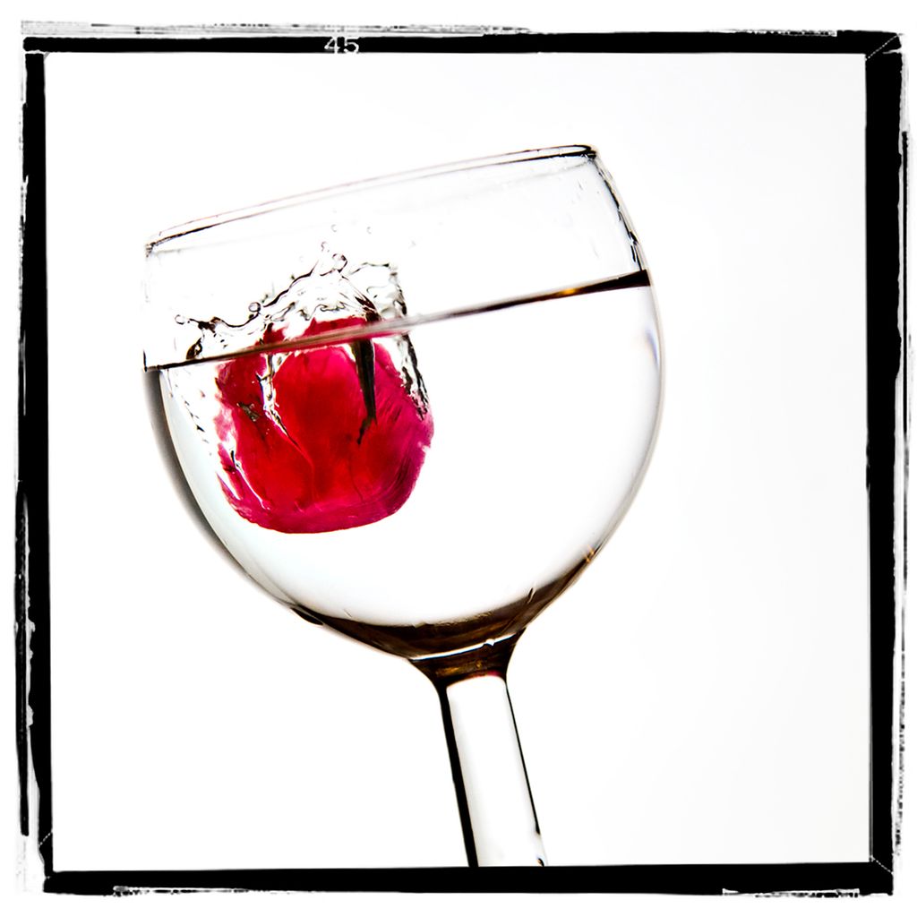 Et rødt drops er kastet oppi et vinglass med klar væske. Lyset framhever dropset. Foto.