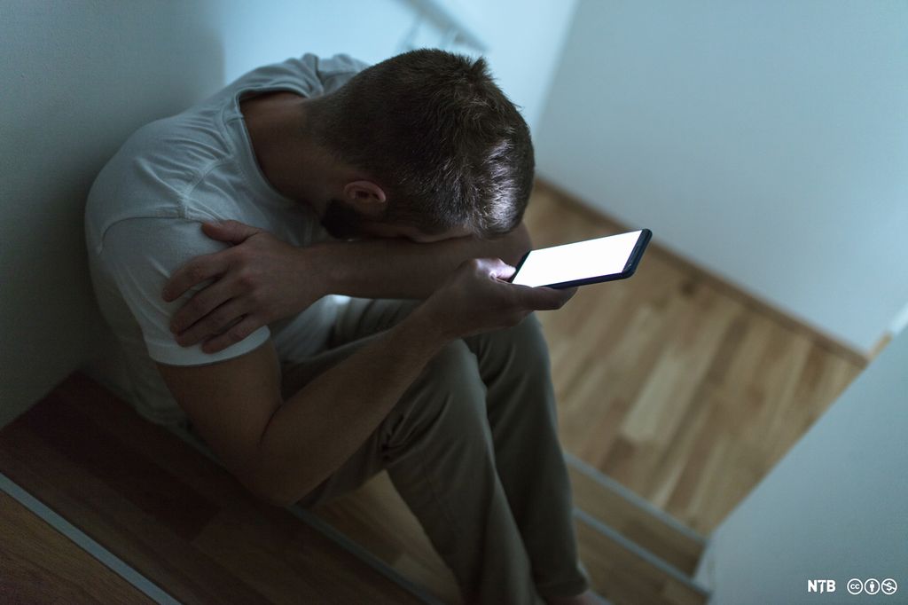 En ung mann sitter med hodet i hendene og holder en mobiltelefon. Foto.