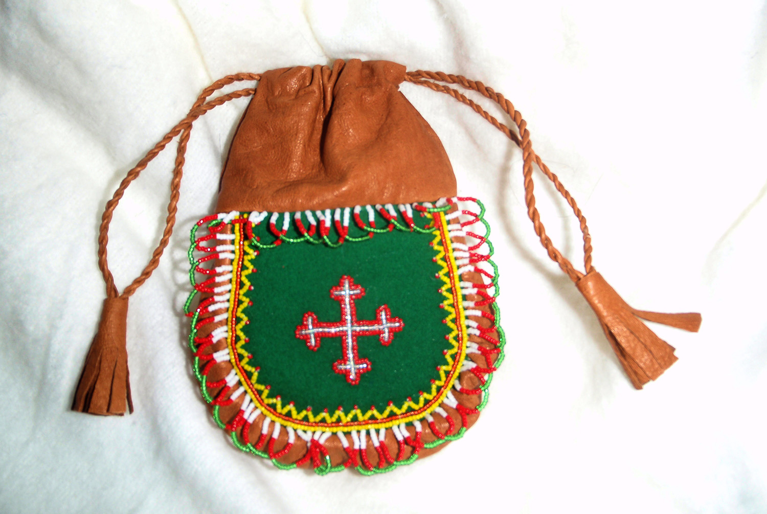 Veska er i brunt skinn med tvinna snøreband. Dekoren er i sterke fargar. I midten eit sølvfarga kors med raud kant på grøn botn. Foto.