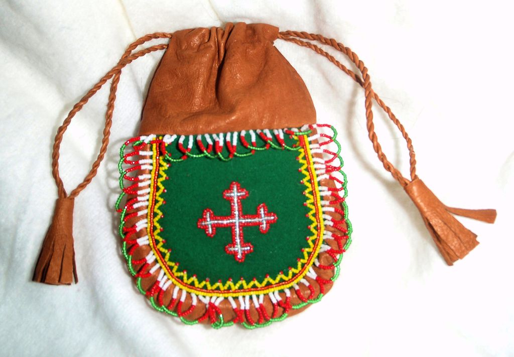 Veska er i brunt skinn med tvinna snøreband. Dekoren er i sterke fargar. I midten eit sølvfarga kors med raud kant på grøn botn. Foto.