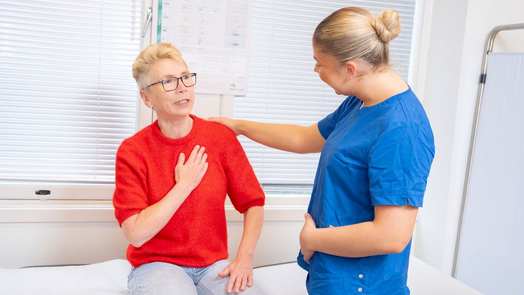 Pasient sitter på undersøkelsesbenk og holder den ene hånda på brystet. Helseservicearbeider holder hånda på skulderen til pasienten og ser på hen. Foto.