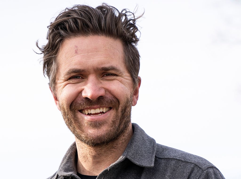 En person smiler til kamera. Han har kort skjegg, rufsete hår, sort t-skjorte, grå skjorte og et stort smil. Foto.