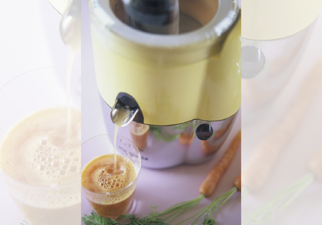 Oransje væske rennet ut av et lite rør fra en stor beholder og ned i et glass. Ved siden av glasset ligger det to gulrøtter. Foto.