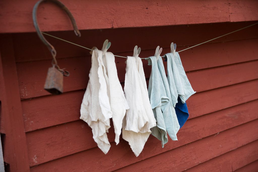 Flere bomullskluter henger til tørk på ei snor. Snora henger langs en rød trevegg, og det henger også ei gammel, rusten bjelle med klave på veggen. Foto.