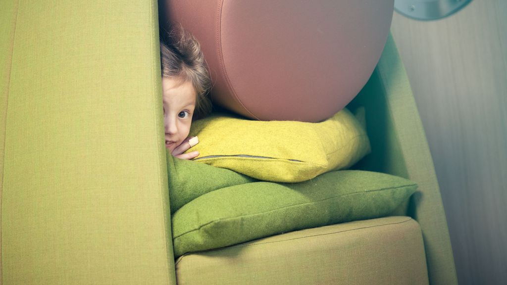 Jente gjemmer seg i ei hytte laget av puter med forskjellige former og farger. Hun titter ut gjennom en liten åpning. Foto.