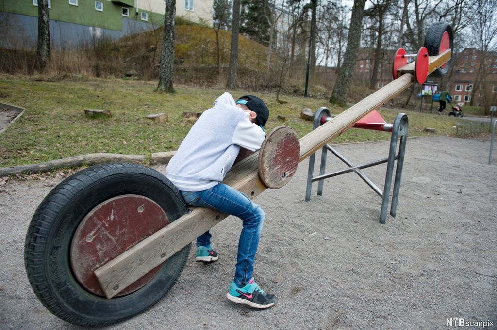 En trist gutt sitter alene på vippehuska på en lekeplass. Han holder armene foran ansiktet. Foto.