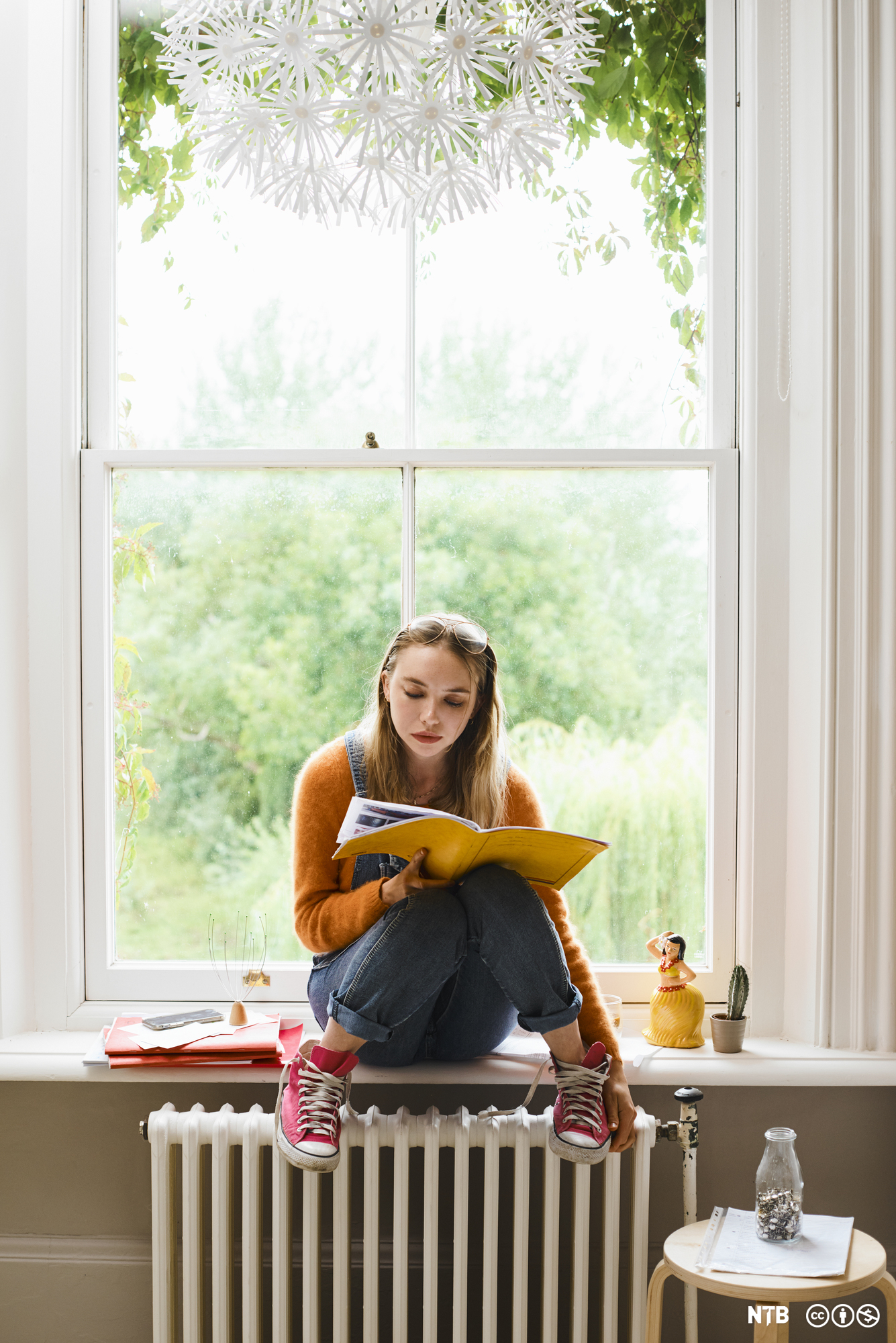 Jente studerer tekst i en bok. Hun sitter i en vinduskarm, har solbriller på panna, løse skolisser og beina på en oljefylt ovn. Foto.