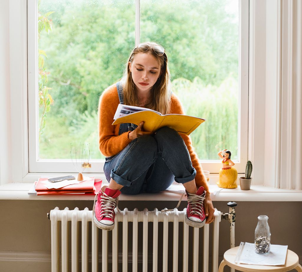 Jente med røde tøysko sitter i vinduskarmen og leser i et hefte med gult omslag. Foto.