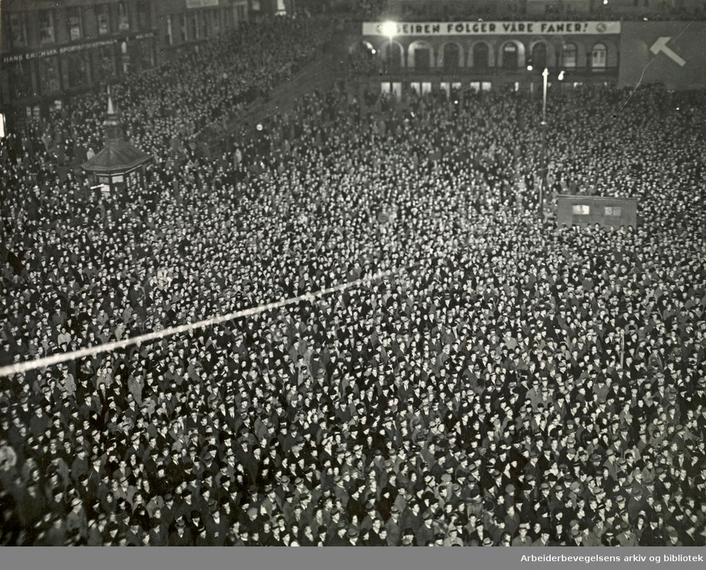Stor folkemasse samlet. På bygget i bakgrunnen står det med store bokstaver: "Seiren følger våre faner!" Svart-hvitt foto.