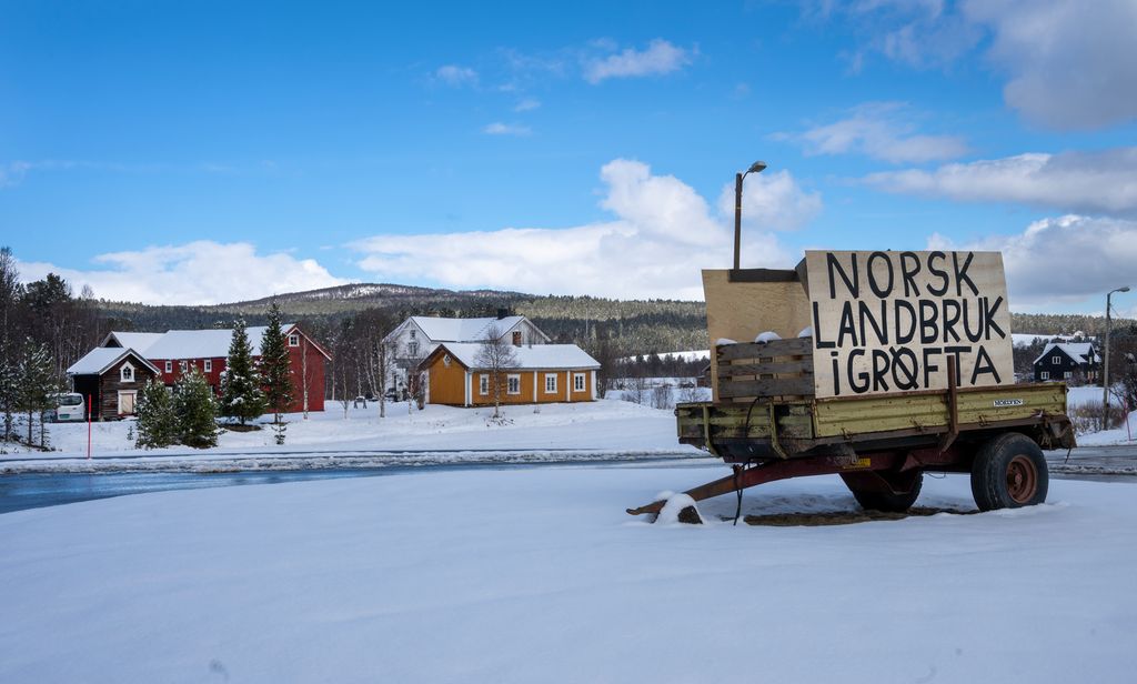 Tilhenger med plakat med teksten Norsk landbruk i grøfta på snødekt jorde foran en gard. Foto.