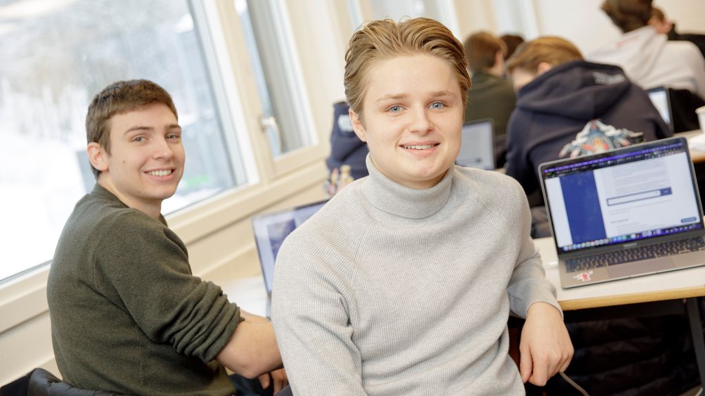 To elever i et klasserom smiler og ser i kameraet. I bakgrunnen er pc-er og medelever. Foto