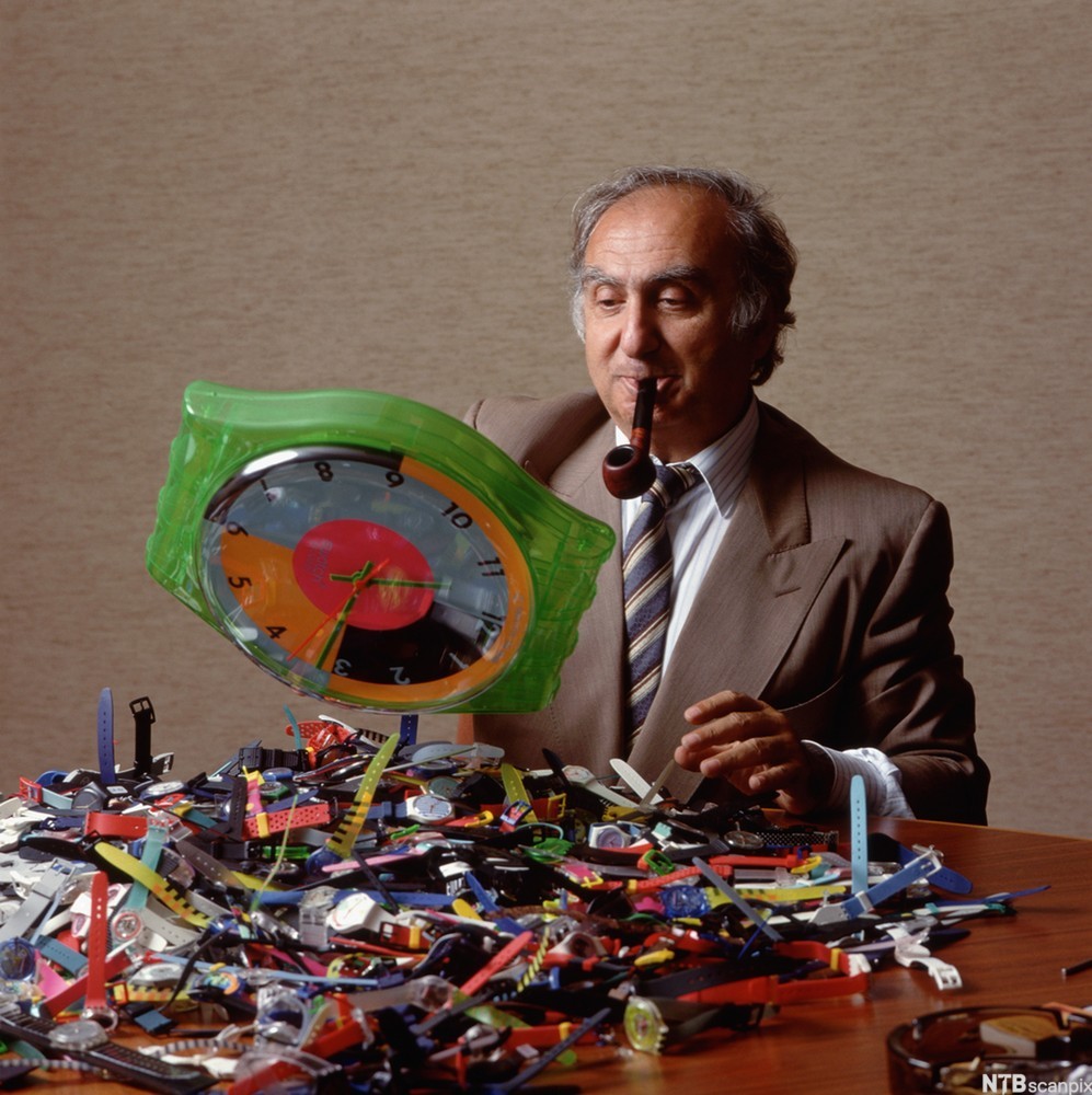 Nicolas Hayek, president i Swatch, sitter ved et bord med mange klokker i en haug foran seg. Foto.