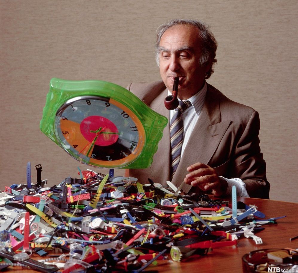 Nicolas Hayek, president i Swatch, sitter ved et bord med mange klokker i en haug foran seg. Foto.