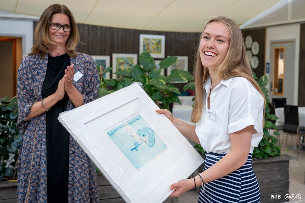 Ei jente smiler mens hun holder et stort bilde. I bakgrunnen klapper en eldre kvinne i hendene. Foto. 