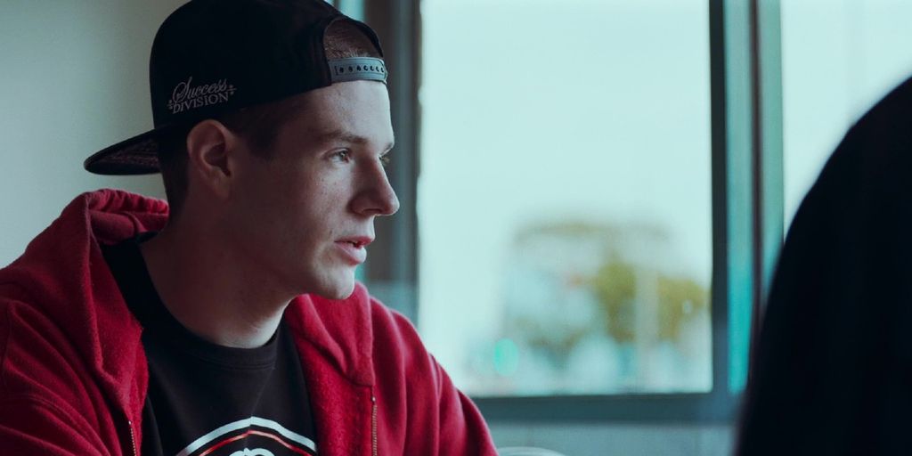 Actor Connor Swindells as the character Adam in the film VS. A teenage boy in a black cap, black t-shirt and a red hoodie. Photo.