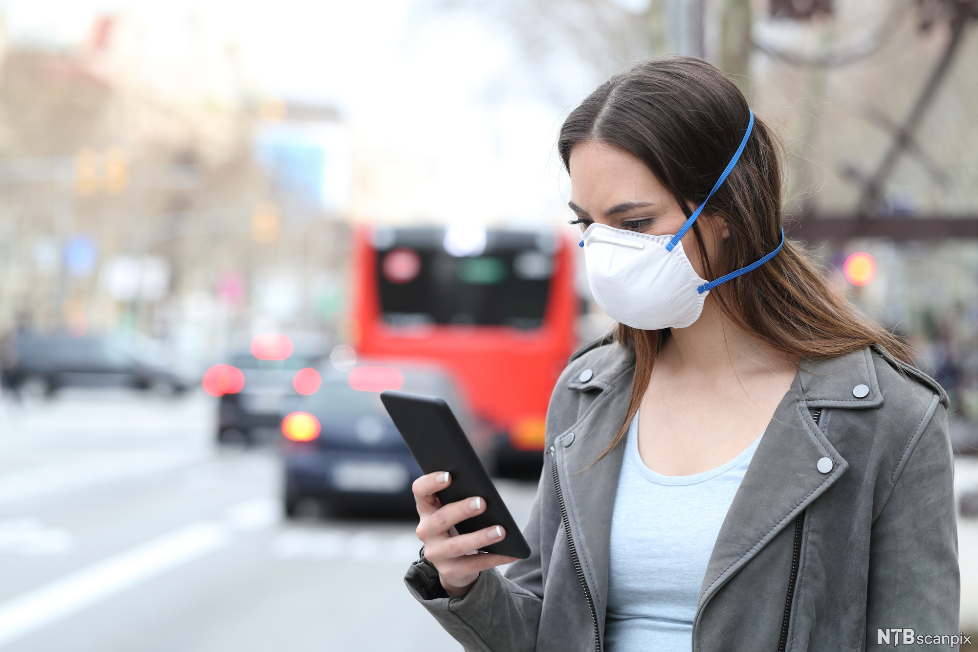 Person med munnbind bruker mobil med bytrafikk i bakgrunnen. Foto.