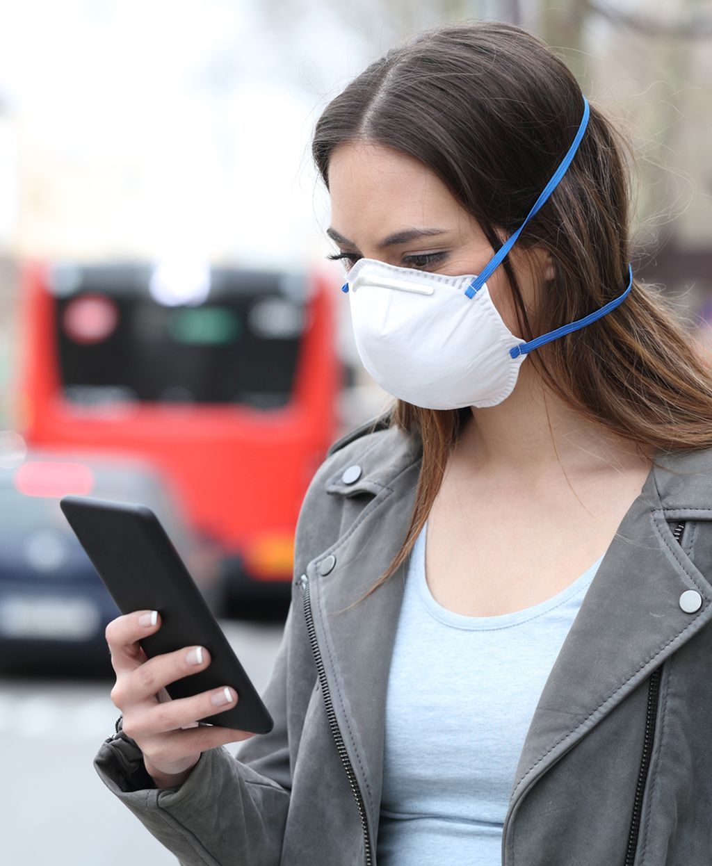 Person med munnbind bruker mobil med bytrafikk i bakgrunnen. Foto.