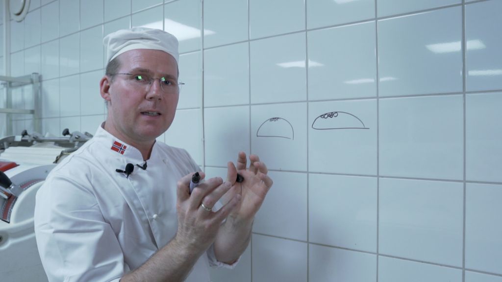 En mann i bakeklær tegner med tusj på flisene på veggen mens han gestikulerer med hendene. Han har tegnet to brød. Foto.