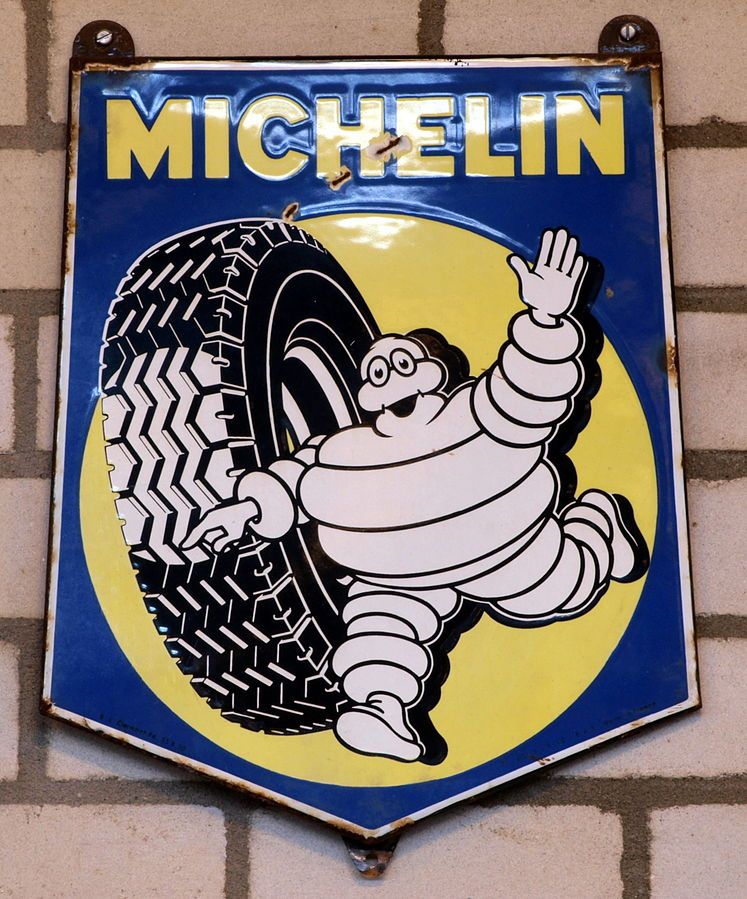 Et metallskilt der det står "Michelin" øverst. Under er det en illustrasjon av et bildekk og en fyldig, hvit figur som smiler og vinker. Foto.