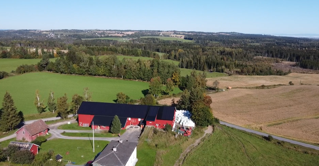 Oversiktsbilde av et gardsbruk med røde driftsbygninger, siloer og et våningshus, omgitt av grønne jorder, skog og åkre. Foto.