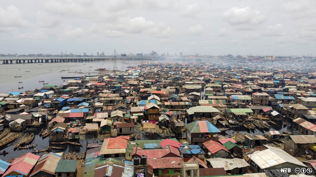 Photo: We see a lot of simply built houses surrounded by water. The houses have roofs in different colours. In the distance we see a line of taller buildings. 