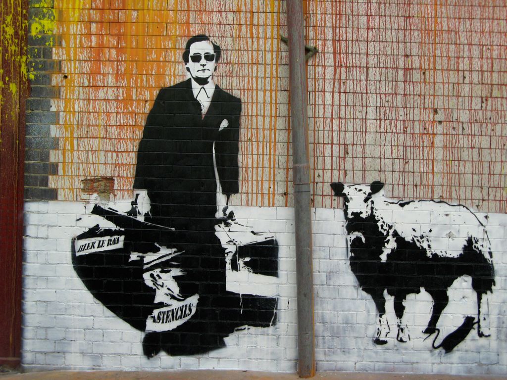 Murgraffiti av en mann i dress og med solbriller som bærer på to vesker. På ei av veskene står det "Blek le rat" på den ene sida og "Stencils" på den andre. Ved siden av mannen er den en sau og ei rotte. Foto.