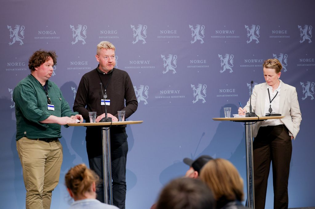 Tre personer presenterer foran ei forsamling. Veggen bak dem er dekket av riksvåpenet og teksten "Norge, departementene" på norsk, samisk og engelsk. Foto.