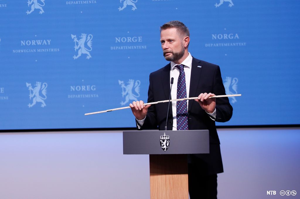 Mann i dress med en tommestokk på en talerstol med riksvåpenet på. Foto.