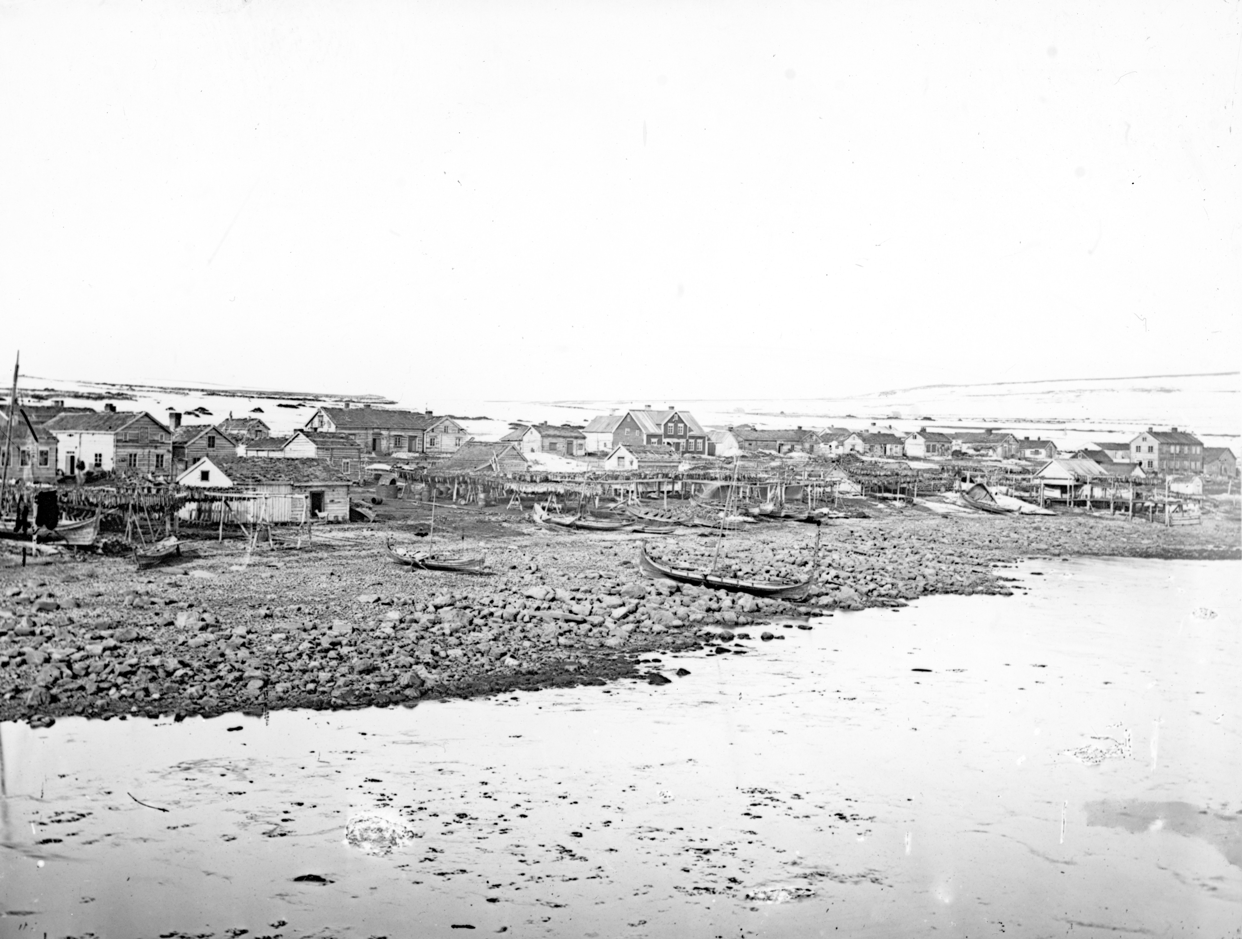 Vadsø på slutten av 1800-tallet. Trehusbebyggelse ved sjøen. Noen få båter i forgrunnen. Foto.